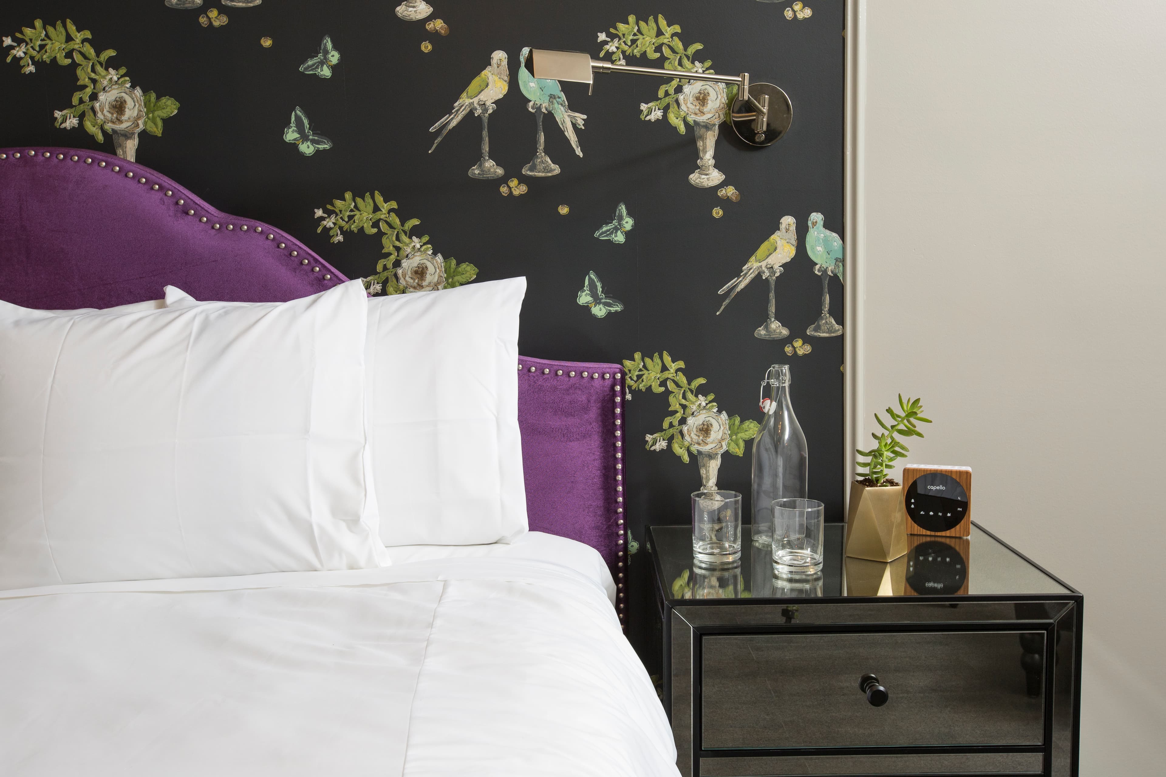 A close up of the pillows, velvet headboard, and nightstand, including glasses of water, succulent plants and a clock radio.