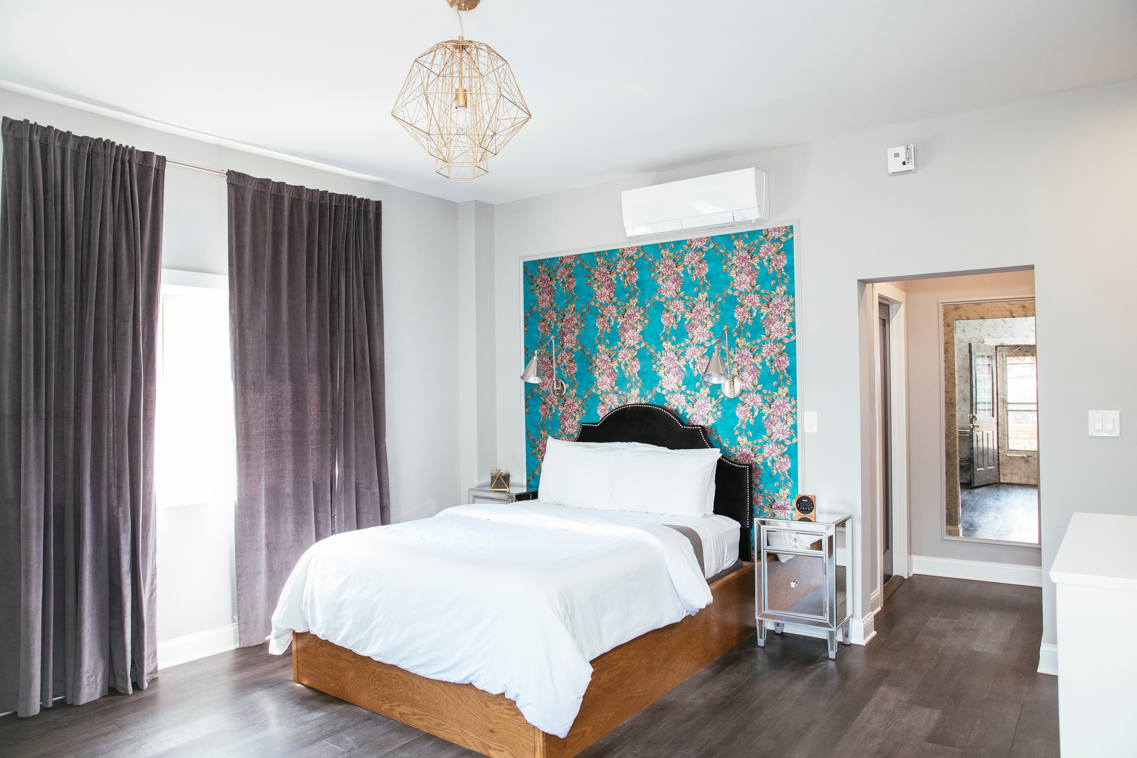 A bright image of Paulina's queen bed, with floral wallpaper, plants on the nightstand and a bottle of water on the bedside table.