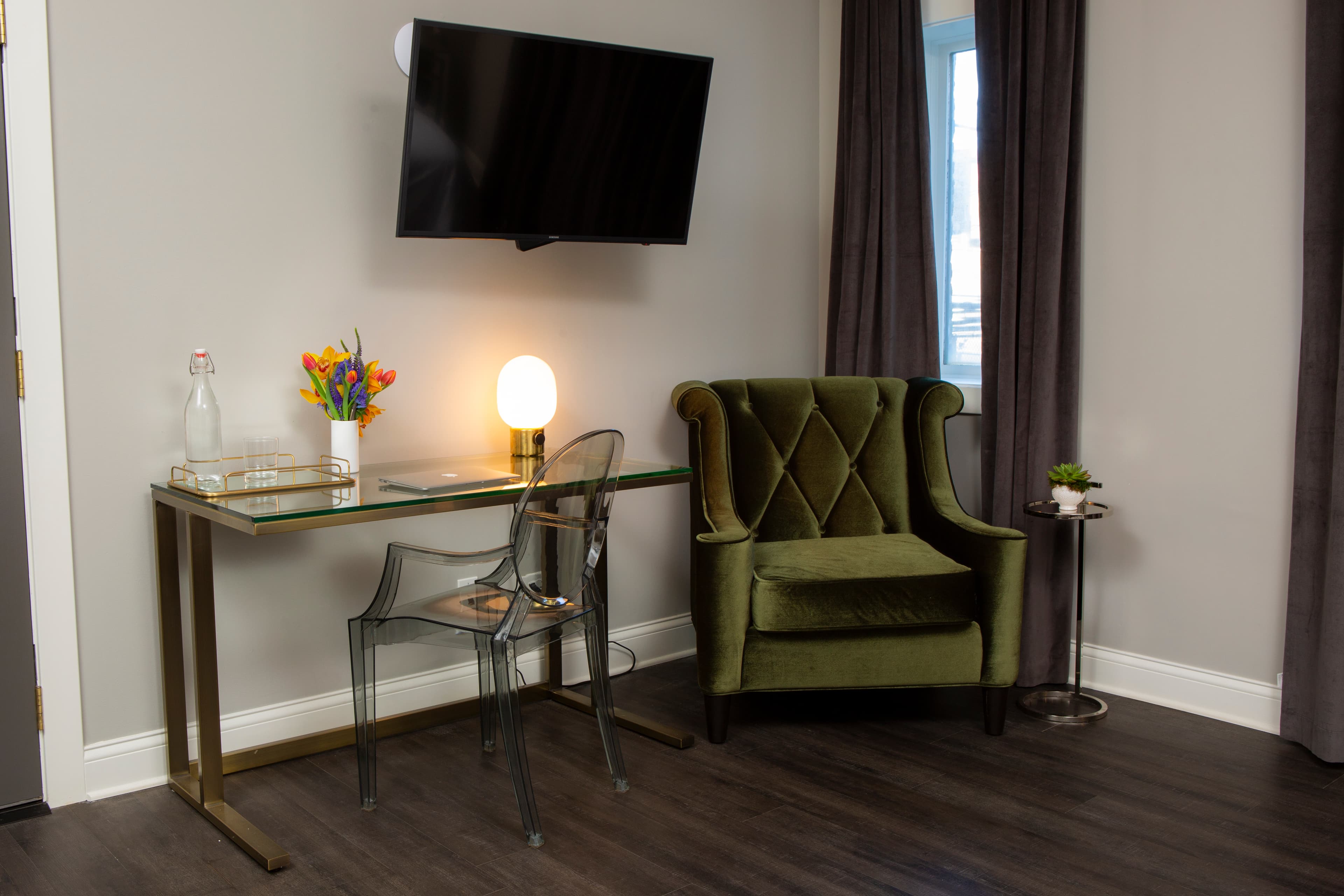 The desk on the other side of Paulina's spacious floor plan, with flowers, a glass bottle of water and a lamp glowing next to a comfortable green velvet armchair by the window.