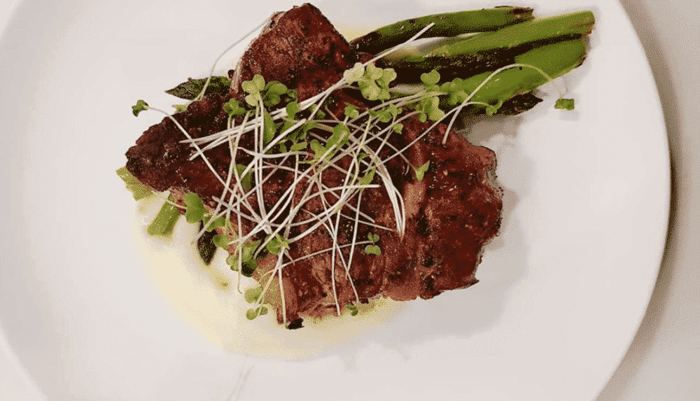A gourmet dish featuring steak garnished with microgreens and served with asparagus on a creamy sauce.