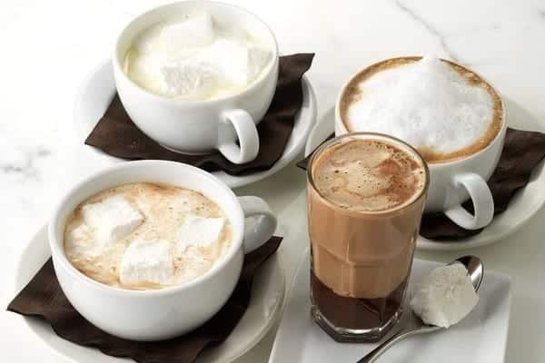 Four cups of beverages with marshmallows on saucers, including a hot chocolate and a cappuccino.