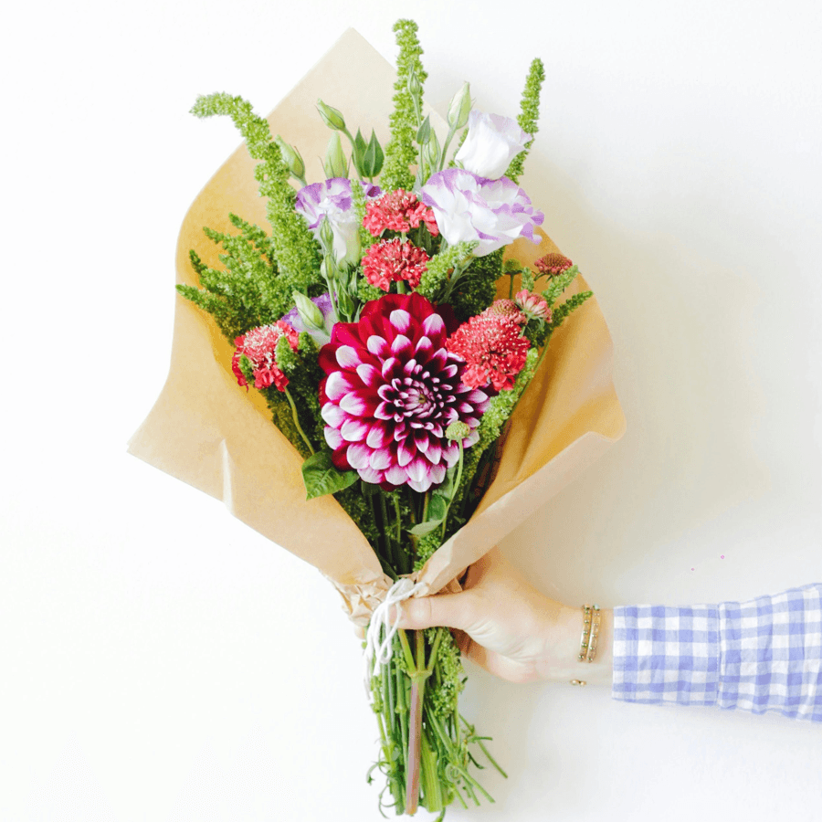 A hand holding a colorful bouquet of flowers wrapped in kraft paper.