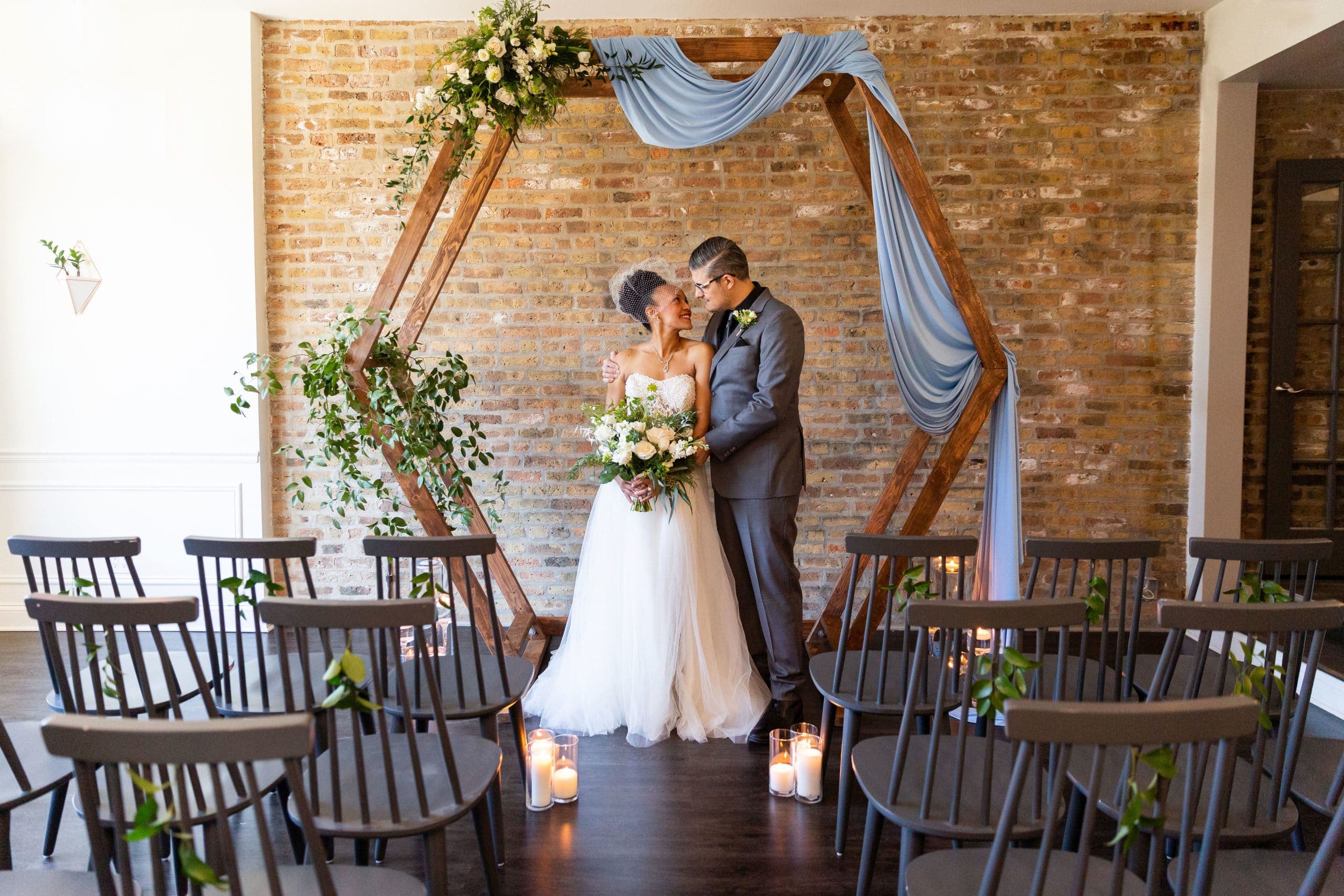 A bride and groom embrace under a floral arch in a wedding ceremony setting.