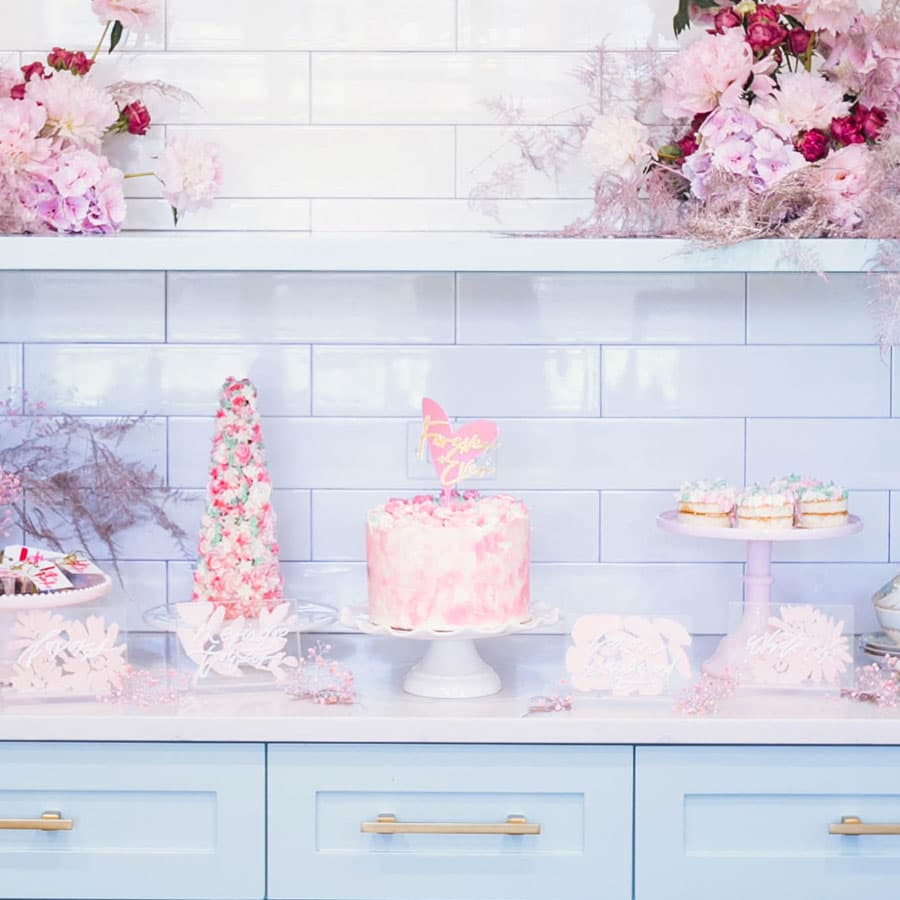A pastel-themed dessert display with a pink cake, colorful treats, and floral decorations on a light blue countertop.