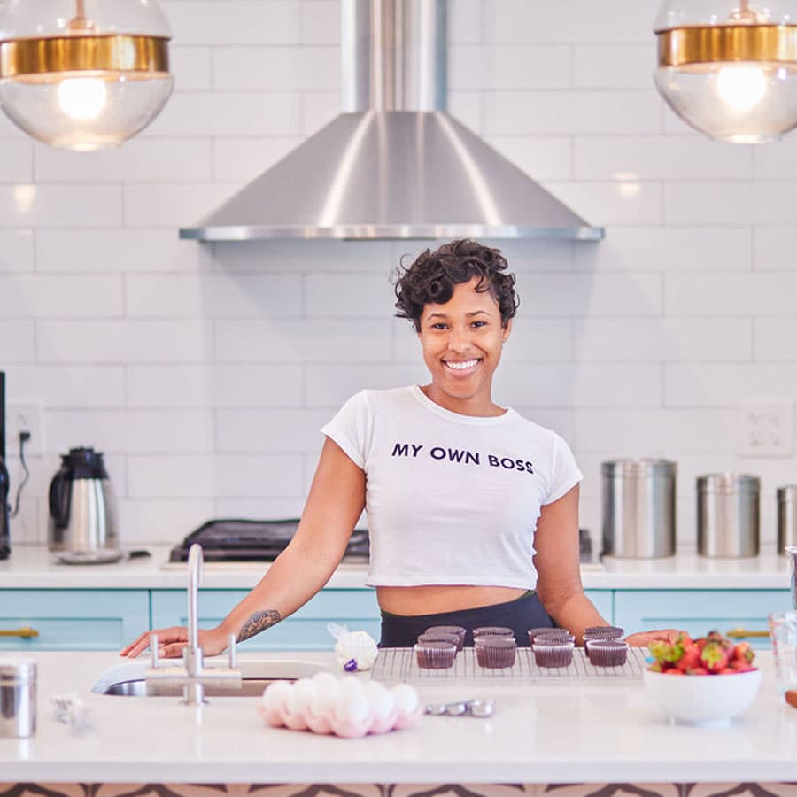 A smiling woman in a kitchen wears a shirt that reads "MY OWN BOSS" while standing next to freshly baked cupcakes.