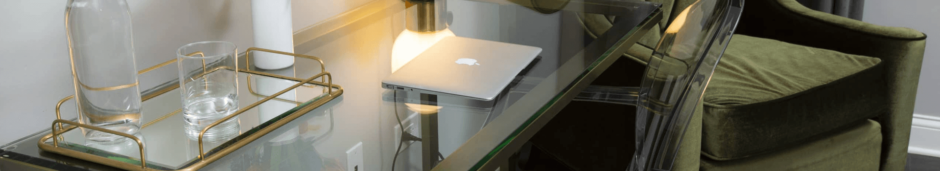 A sleek glass desk with a laptop, a watering can, and a green armchair.