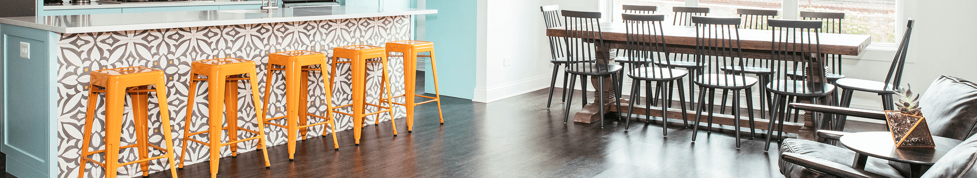 A modern kitchen area featuring orange bar stools, a patterned backsplash, and a large dining table surrounded by black chairs.