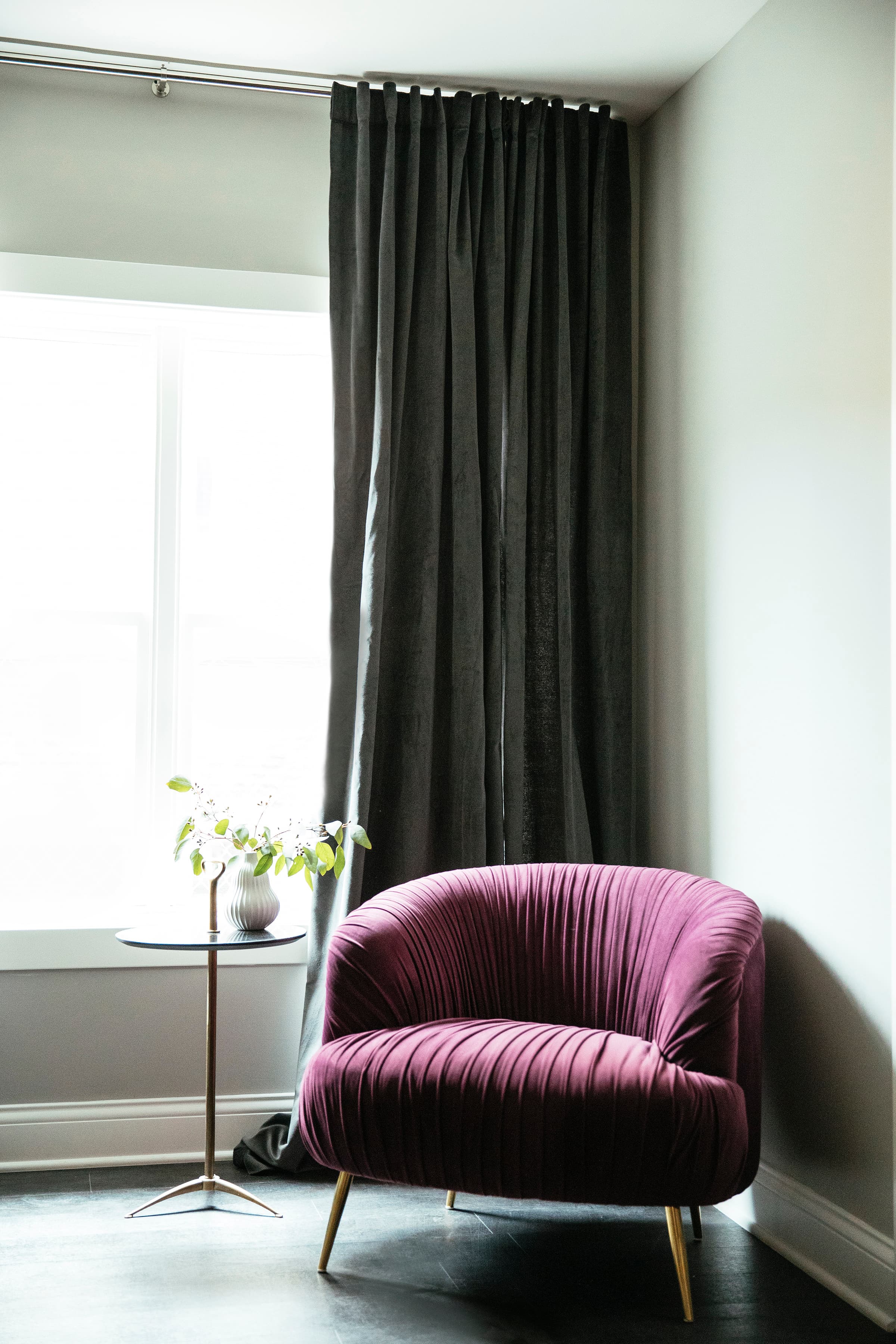 The soft floor length curtains over the window, a red velvet armchair with flowers on a stand beside it, and a desk with glasses of water.
