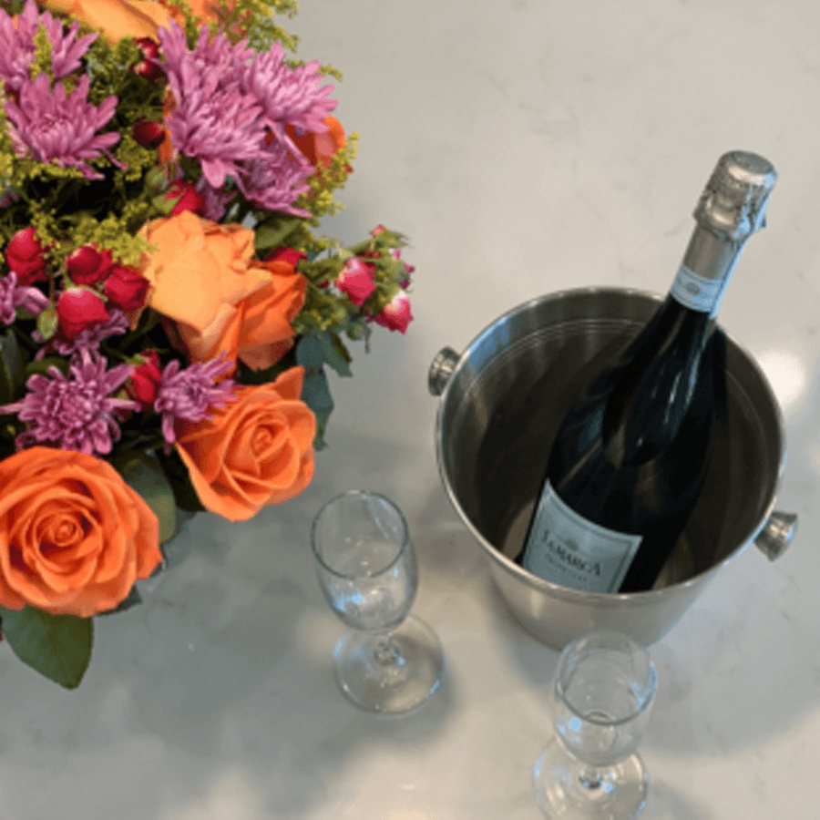 A bouquet of colorful flowers beside a bottle of champagne in an ice bucket and two glasses.