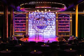 A brightly lit comedy stage with a microphone and colorful backdrop in a cozy venue.
