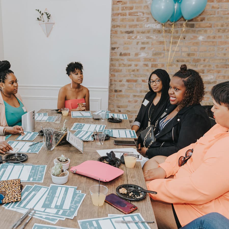 A group of women engages in conversation around a table adorned with notes and refreshments.