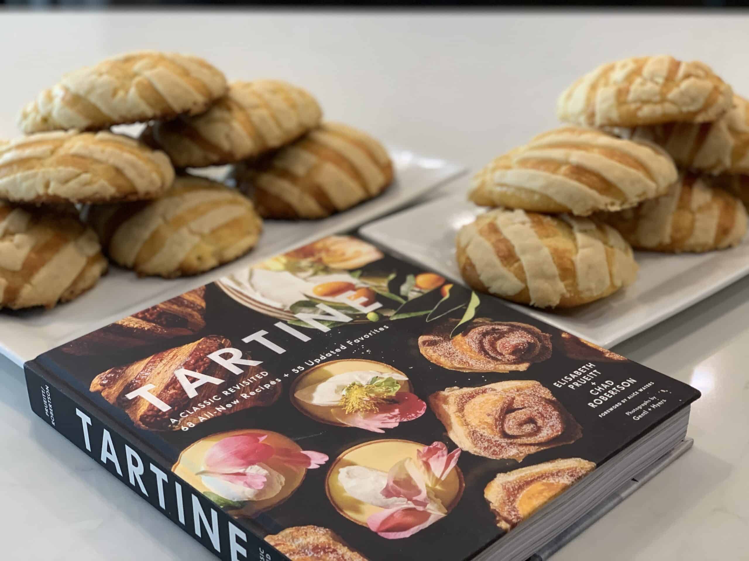 A cookbook titled "Tartine" lies open beside a platter of freshly baked bread rolls. A cookbook titled "Tartine" lies open beside a platter of freshly baked bread rolls.