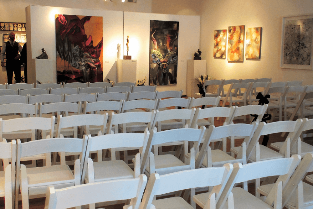Empty white chairs arranged in rows in an art gallery featuring colorful paintings and sculptures.