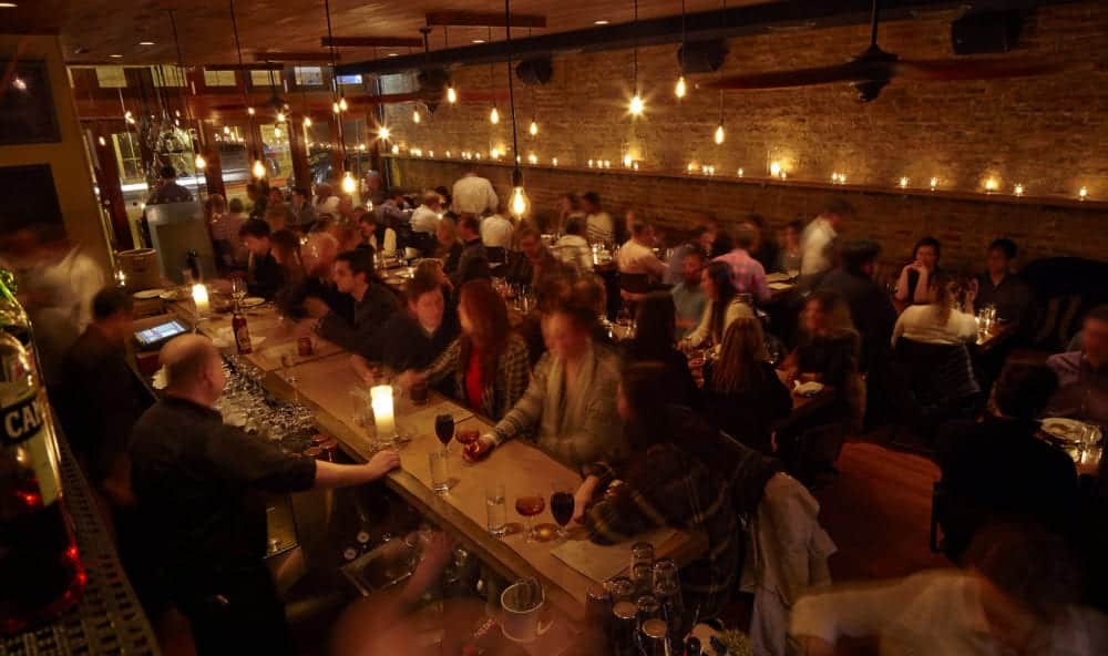 A bustling restaurant scene with dim lighting and patrons enjoying drinks and meals at the bar and tables. A bustling restaurant scene with dim lighting and patrons enjoying drinks and meals at the bar and tables.