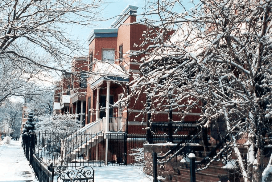 A row of brick houses surrounded by snow-covered trees and a white fence.