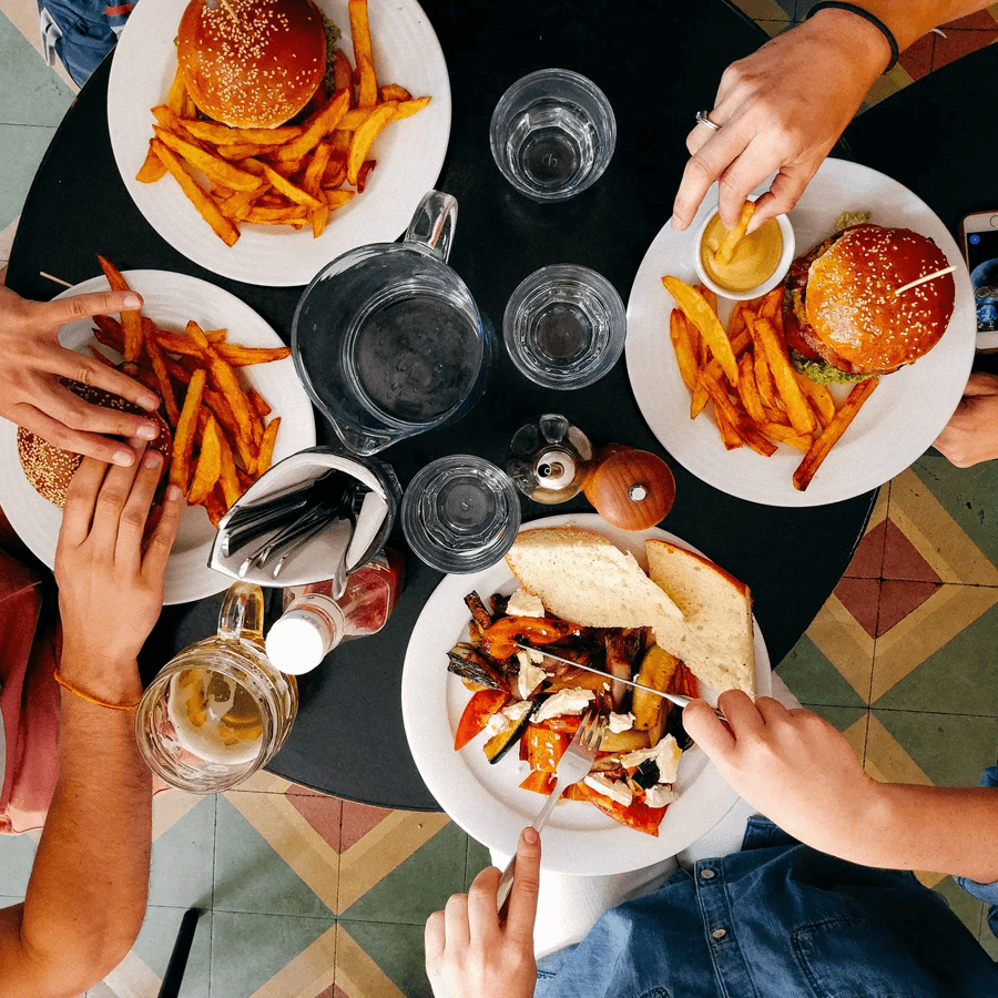 A bird's-eye view of a table set with burgers, fries, and a colorful vegetable dish, surrounded by hands reaching for the food.