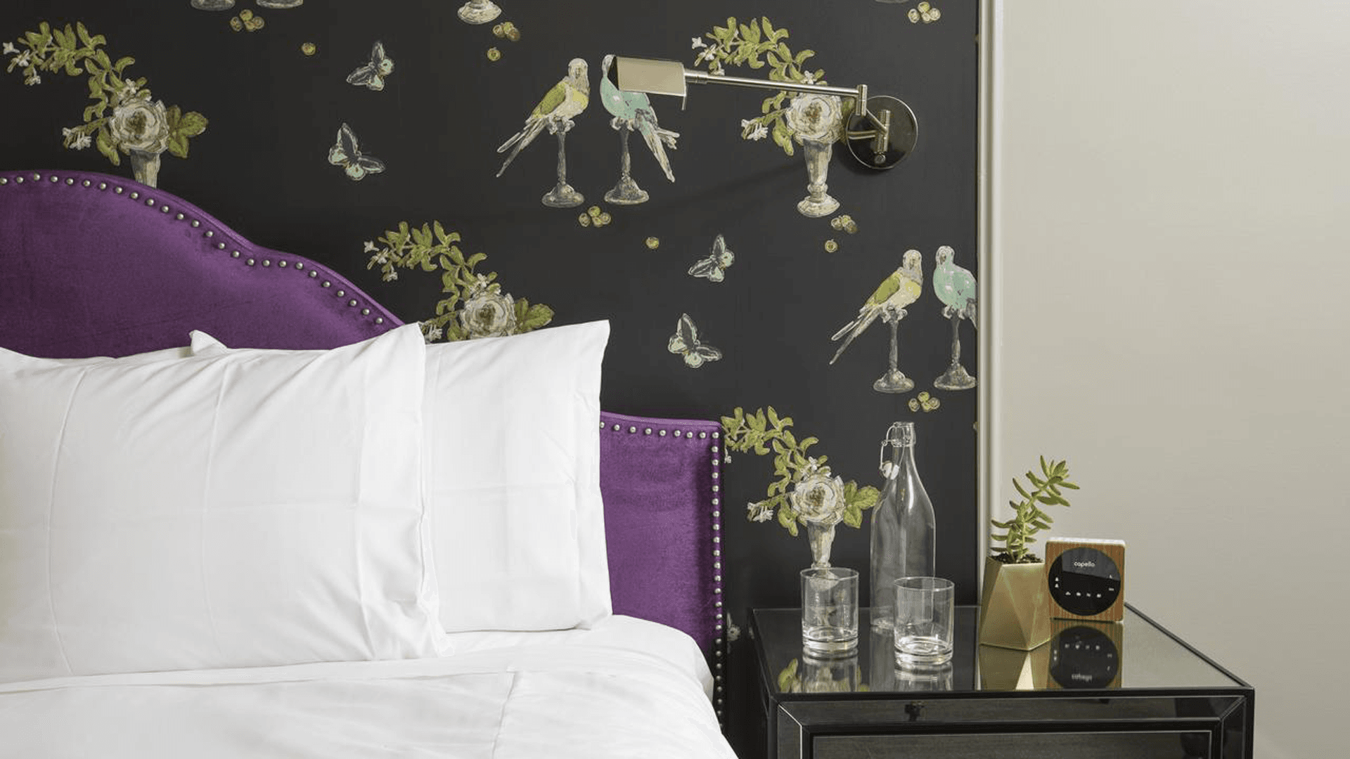 A stylish bedroom featuring a purple upholstered headboard, white pillows, and a wall with floral and bird patterns.