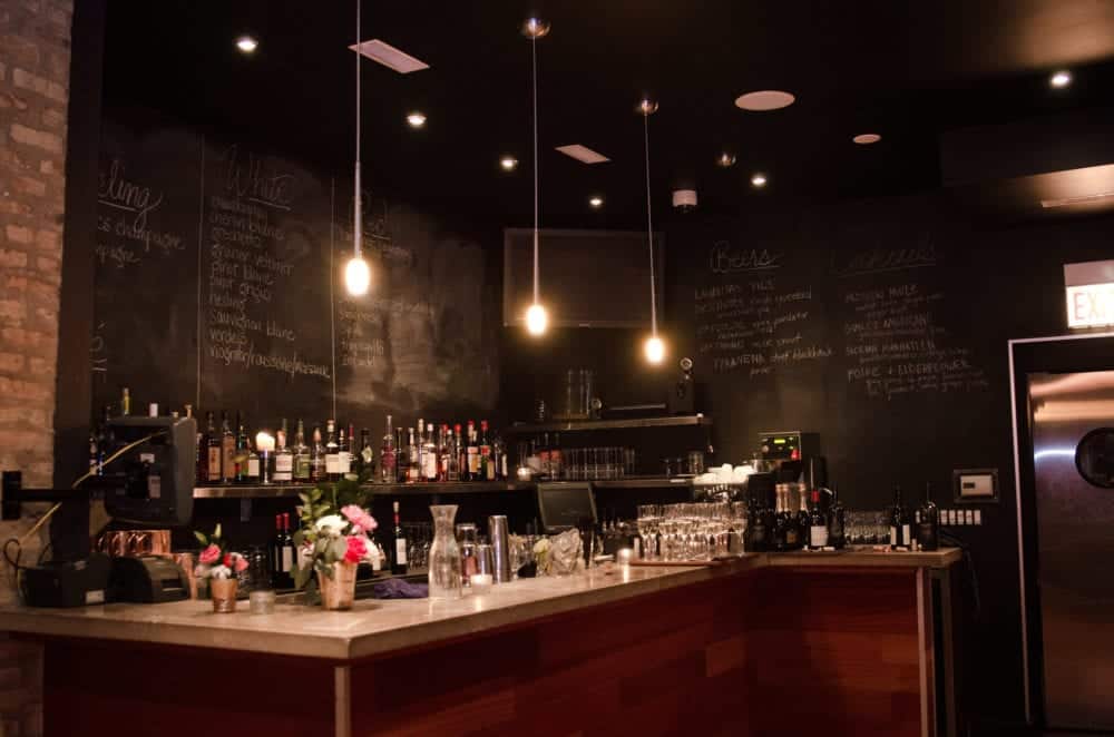 A dimly lit bar with a chalkboard menu, a variety of bottles on the shelves, and glassware arranged neatly on the counter.