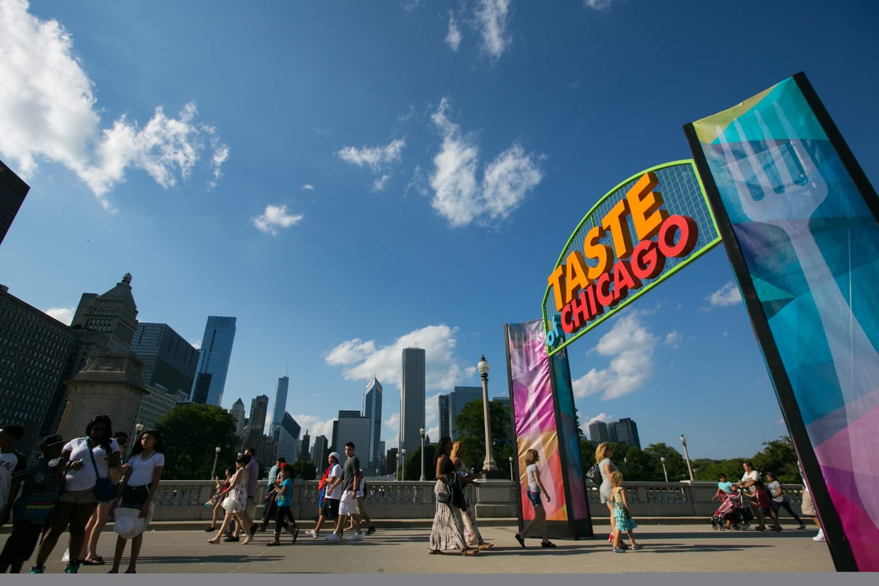 A vibrant sign for "Taste of Chicago" welcomes visitors under a blue sky with clouds and city skyscrapers in the background.