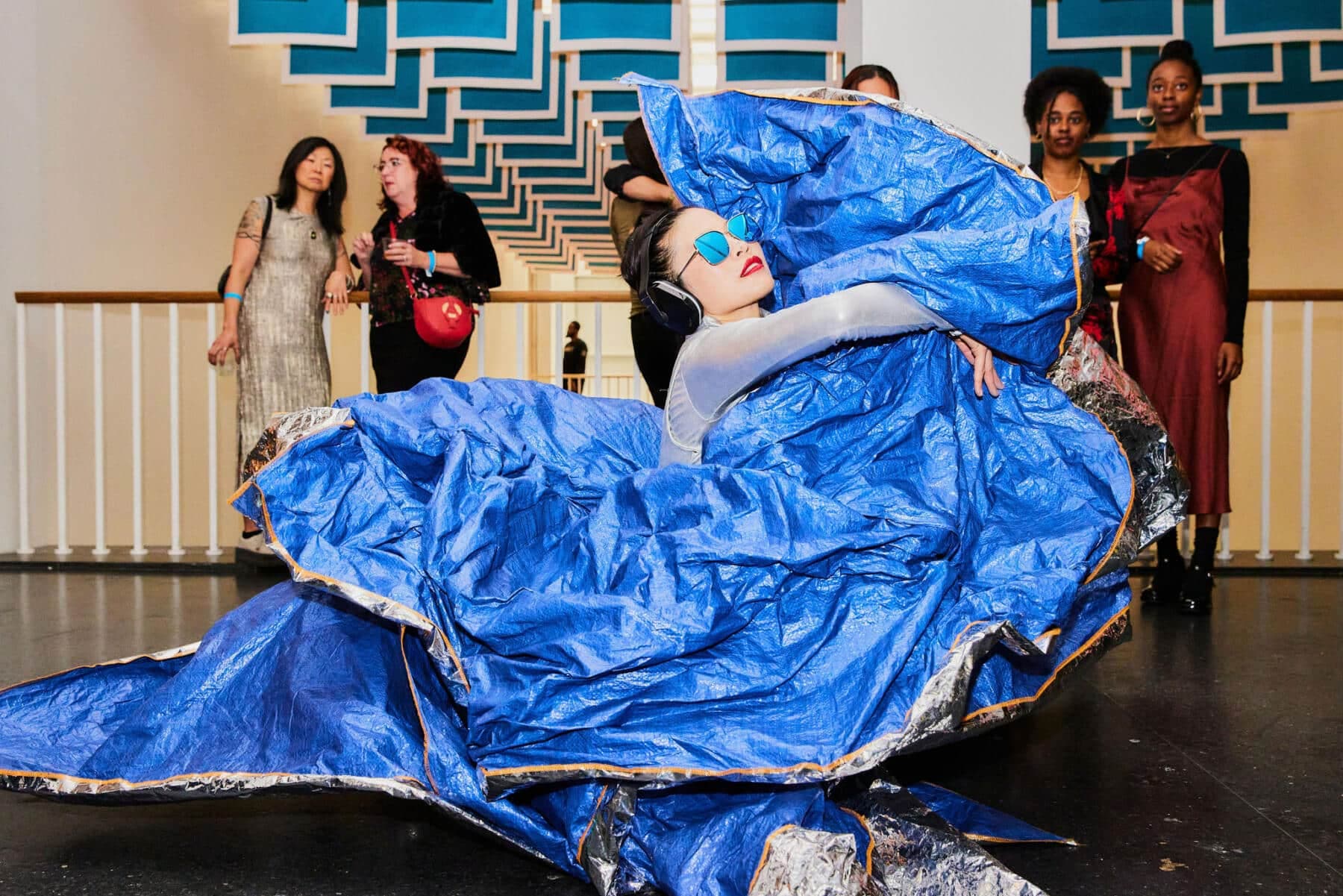 A performer in a blue costume poses dramatically on stage, surrounded by onlookers in the background. A performer in a blue costume poses dramatically on stage, surrounded by onlookers in the background.