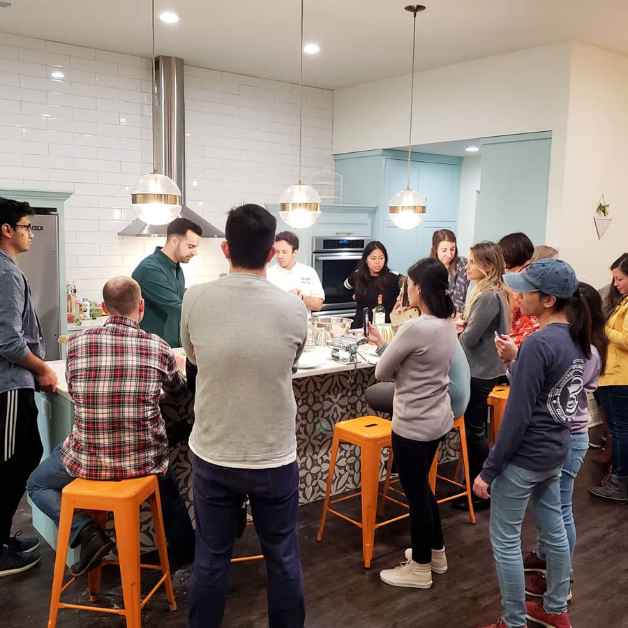 A group of people gathers in a modern kitchen, attentively watching a cooking demonstration.