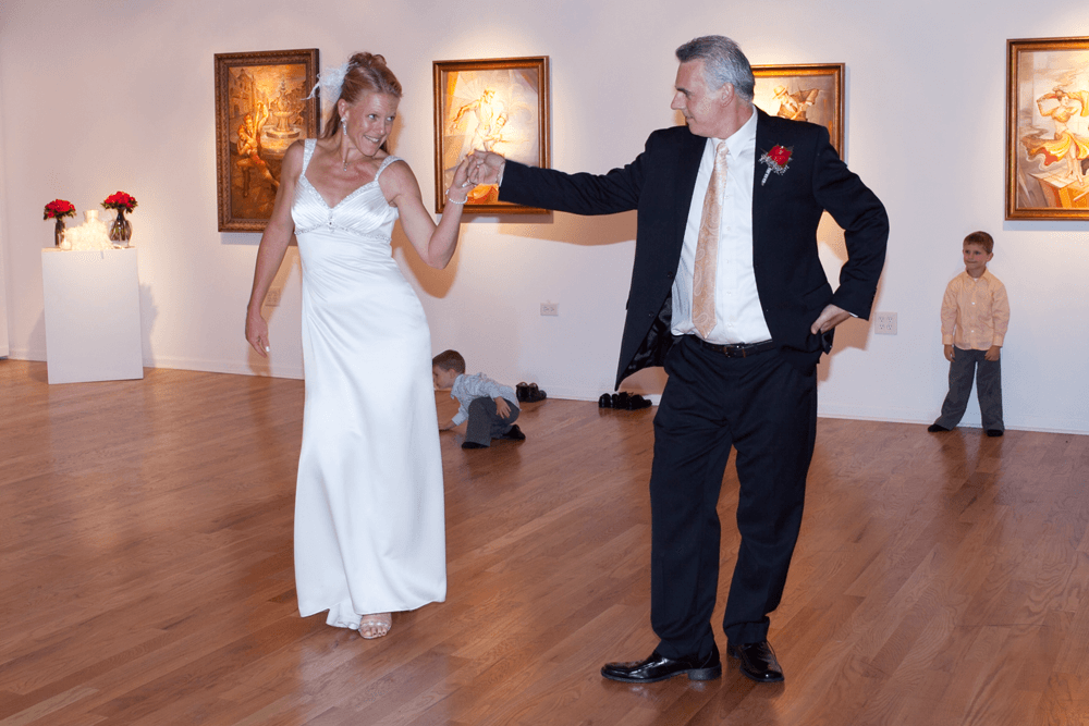 A bride in a white gown dances with a man in a suit in an art gallery, while two young boys watch nearby.