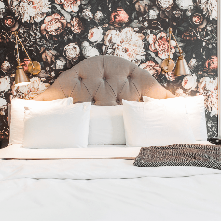 A neatly made bed with white linens and a tufted headboard against a floral wallpaper backdrop.