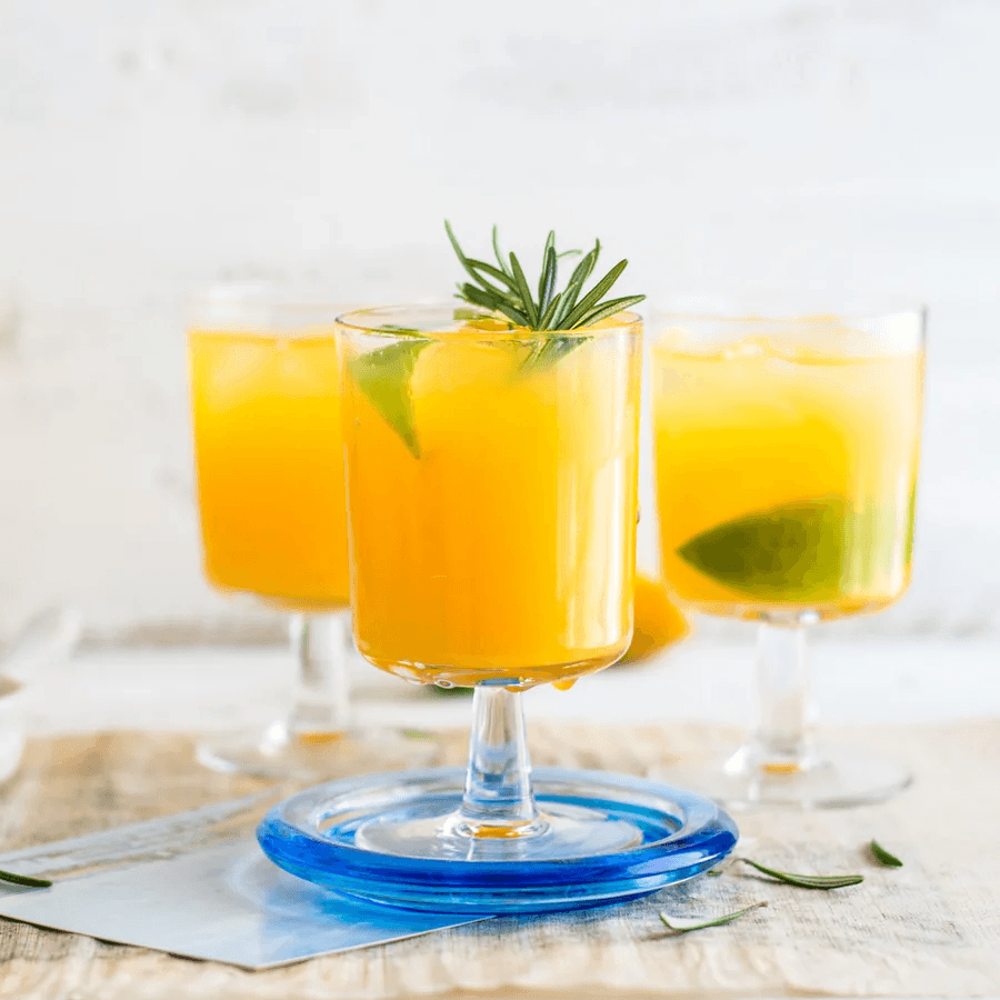 Three glasses of yellow drinks garnished with lime and rosemary on a wooden surface.