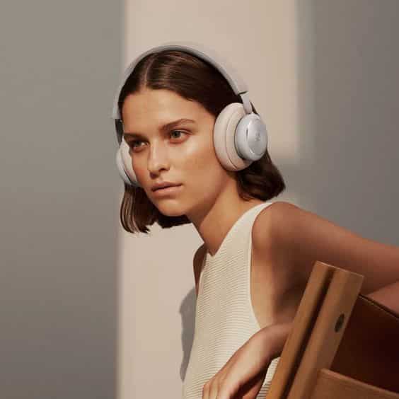 A woman with short hair wearing large headphones leans against a wooden chair in a softly lit environment. A woman with short hair wearing large headphones leans against a wooden chair in a softly lit environment.