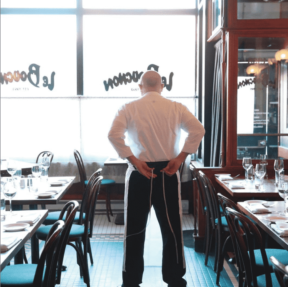 A chef stands with hands behind his back in an empty, elegantly set restaurant.