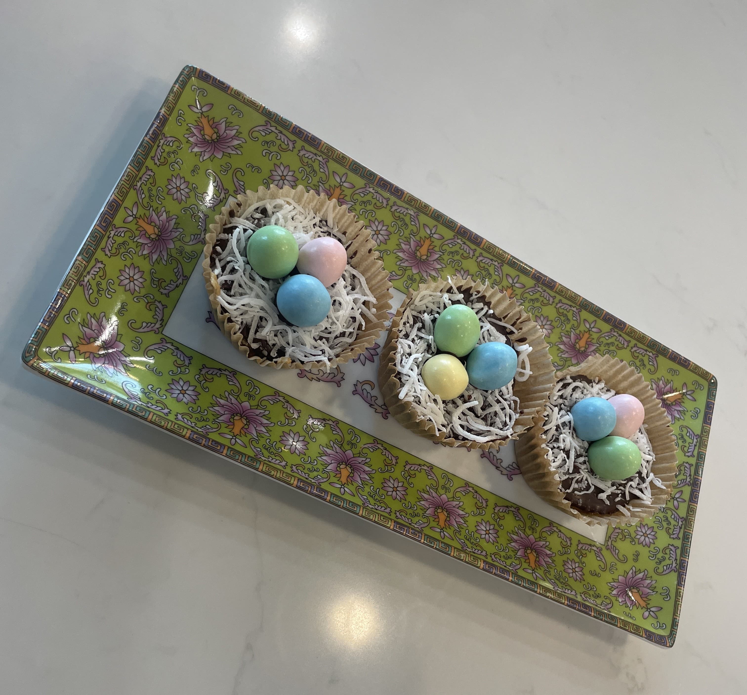 Three dessert nests made of chocolate and coconut, topped with colorful candy eggs, on a decorative plate.
